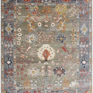 May include: A rectangular area rug with an intricate floral design. The rug features a central field of muted green with a border of blue and red. The floral patterns include shades of red, yellow, and white, creating a detailed and ornate appearance.