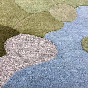 May include: A close-up of a rug with a landscape design. The rug features a river of light blue, surrounded by green and gray organic shapes, creating a textured, plush surface. The design evokes a natural, flowing aesthetic.