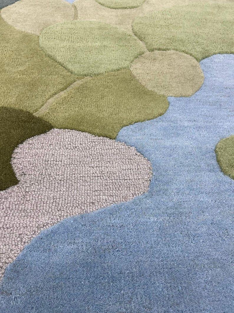 May include: A close-up of a rug with a landscape design. The rug features a river of light blue, surrounded by green and gray organic shapes, creating a textured, plush surface. The design evokes a natural, flowing aesthetic.