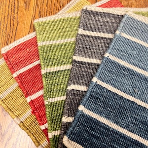 May include: A collection of colorful, rectangular woven placemats. Each mat features horizontal stripes in shades of yellow, red, green, gray, and blue, with white stripes. The mats are arranged in a fan shape, showcasing their varied hues and textures.