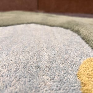 May include: Close-up of a soft, textured rug with a light blue, green, and yellow color scheme. The rug has a plush, looped pile and a whimsical, abstract design. The colors are muted and create a calming aesthetic.