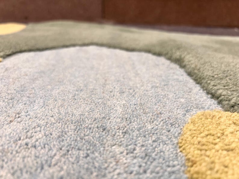 May include: Close-up of a soft, textured rug with a light blue, green, and yellow color scheme. The rug has a plush, looped pile and a whimsical, abstract design. The colors are muted and create a calming aesthetic.