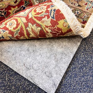 May include: A close-up of a gray rug pad with a patterned rug folded over it. The rug has a red, yellow, and blue design with a fringe border.