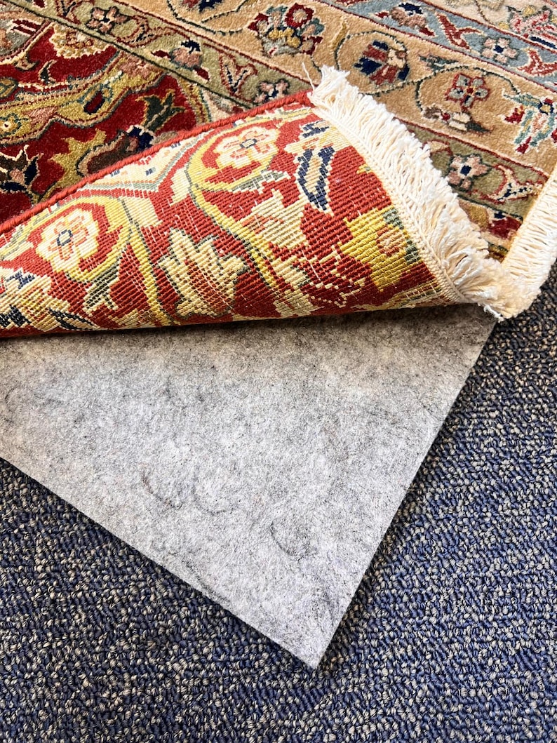 May include: A close-up of a gray rug pad with a patterned rug folded over it. The rug has a red, yellow, and blue design with a fringe border.