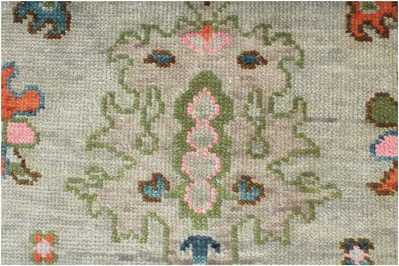 May include: Close-up of a light gray rug with a detailed floral pattern. The design features a central motif with pink, green, and brown elements, surrounded by stylized floral shapes in orange, blue, and green. The rug has a textured appearance.