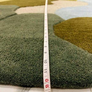 May include: A close-up of a green, blue, and tan area rug with an abstract design. A measuring tape shows the rug's thickness. The rug has a plush, textured surface and a landscape-like pattern.