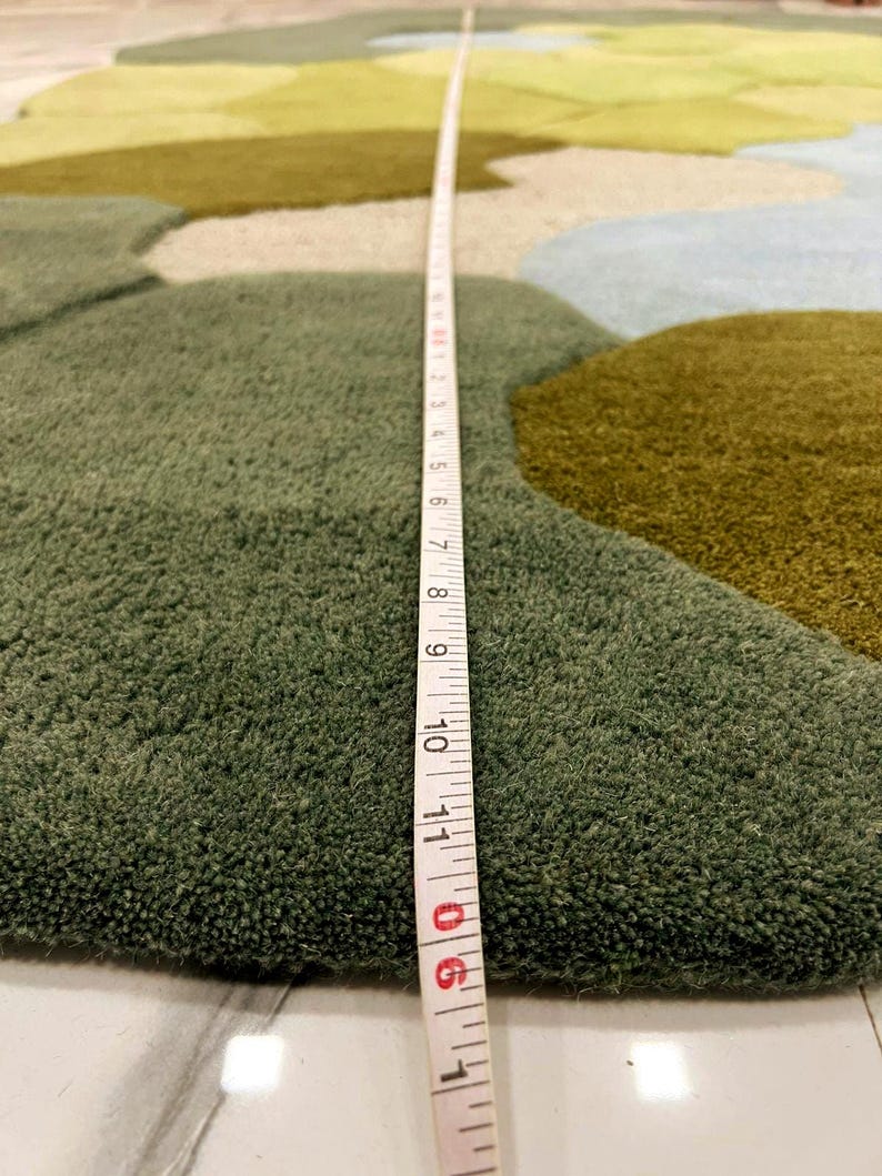 May include: A close-up of a green, blue, and tan area rug with an abstract design. A measuring tape shows the rug's thickness. The rug has a plush, textured surface and a landscape-like pattern.