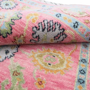 May include: A close-up of a pink rug with a detailed floral pattern. The rug features a combination of colors, including pink, blue, yellow, and green. The design includes floral and geometric elements.