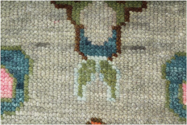 May include: Close-up of a textured rug with a repeating floral pattern. The rug features a light gray background with accents of teal, pink, green, and brown. The design is symmetrical, with the floral elements evenly spaced across the surface.