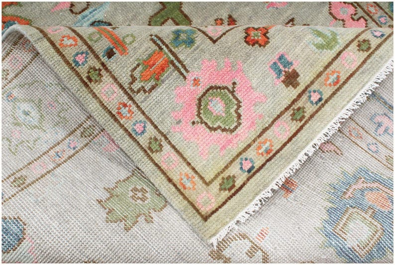 A close-up of a light green rug with a pink and white floral pattern. The rug has a fringe border and is folded over on itself. The rug is made of a soft, woven material.