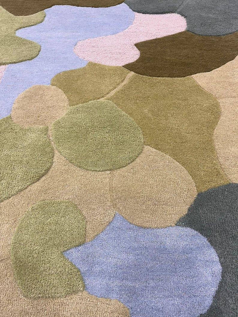 May include: A colorful, abstract rug featuring a pattern of organic shapes in various shades. The rug incorporates hues of green, brown, blue, and pink, creating a soft, textured surface. The design is reminiscent of a landscape or a map.