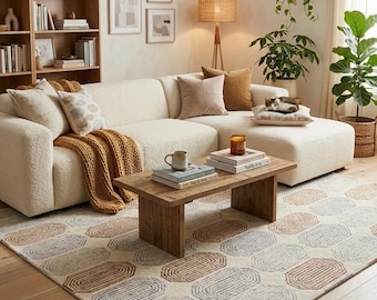 May include: A cozy living room scene featuring a cream-colored sectional sofa, a wooden coffee table, and a patterned rug. The rug has an off-white background with brown, blue, and rust-colored oval designs. Decorative pillows and a mustard-colored throw blanket add warmth.