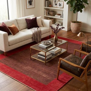 May include: A living room scene featuring a cream-colored sofa with burgundy and orange pillows, a glass-top coffee table, and two leather armchairs. A large red rug with a darker border covers the floor, complementing the room's warm tones.