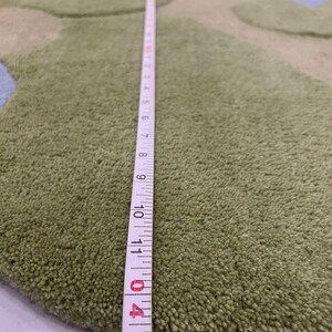 May include: A green, round rug with a textured surface and a design of overlapping shapes in shades of green and blue. A measuring tape shows a length of approximately 11 inches.