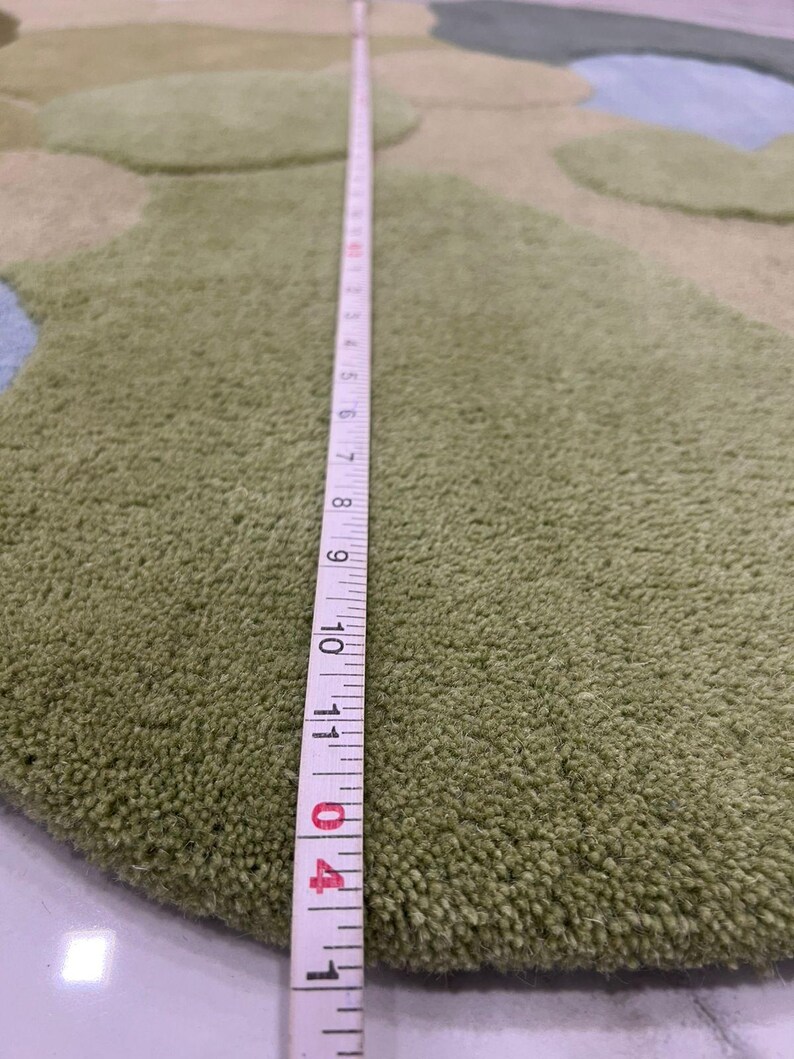 May include: A green, round rug with a textured surface and a design of overlapping shapes in shades of green and blue. A measuring tape shows a length of approximately 11 inches.