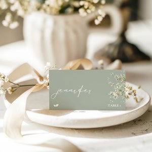 May include: A light green place card with white lettering that reads "jennifer" and "TABLE 1". The card is decorated with a watercolor floral design and is sitting on a white plate.
