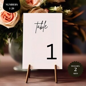 May include: A white table number card with the word "table" in cursive and the number "1" in a bold font. The card is propped up on a wooden stand. The image includes text that says "Numbers 1-20" and "Includes 2 Sizes".