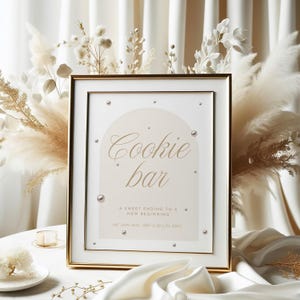 May include: A framed sign with gold trim and the words "Cookie bar" in elegant script. The sign includes the text "A sweet ending to a new beginning" and is decorated with faux pearls. The background features pampas grass and white fabric.