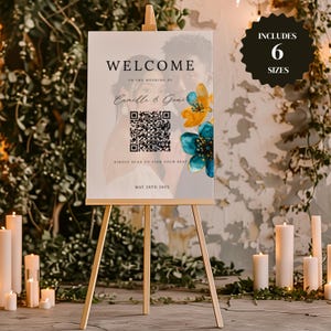 以下が含まれることがあります： 花柄とQRコードがデザインされた結婚式用のウェルカムサイン。サインには「Camille & Ganeの結婚式へようこそ」と「席を探すにはスキャンしてください」と書かれています。日付は2025年5月28日です。