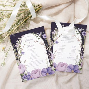 May include: Two wedding ceremony programs with floral designs. The programs have a dark blue background with a white arch and floral accents in purple and lavender. The programs are on a cream-coloured surface with a bouquet of flowers and a white ribbon.