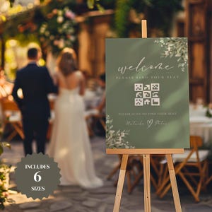 以下が含まれることがあります： 白い花のアクセントとQRコードが付いた緑色のウェルカムサイン。サインには「ようこそ、席を見つけてください。席を見つけるにはスキャンしてください。ナターシャ＆ピーター」と書いてあります。