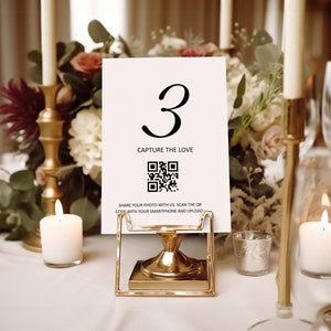 May include: White card with the number 3 and the text "Capture the Love" printed on it. A black and white QR code is below the text. The card is on a gold metal stand.