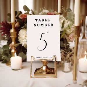 May include: A white card with black text reading "Table Number 5" and "Sabrina & Felix" on a gold metal stand. The card is surrounded by candles and flowers.