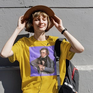 May include: A person wearing a yellow t-shirt with a purple square print featuring a portrait of a man with a mustache and a dark blue jacket. The person is wearing a brown hat and has a black backpack.