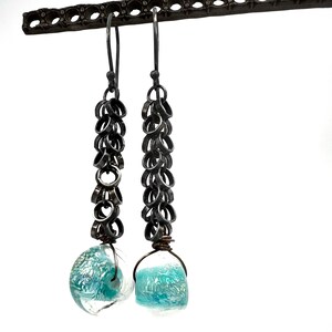 May include: A pair of dangle earrings with a chainmail design. The earrings feature a series of small, dark metal rings connected to a clear glass bead with turquoise and white swirls. The earrings are suspended from a dark metal hook.