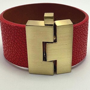May include: A red leather cuff bracelet with a textured surface and a gold-colored metal clasp. The bracelet is wide and has a rectangular clasp that fastens in the center. The interior of the bracelet is a lighter brown color.