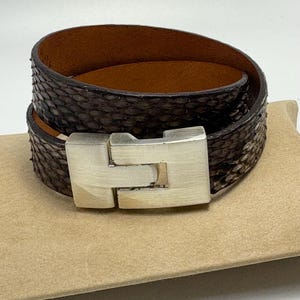 May include: A wrap-around bracelet with a dark brown snakeskin pattern. The bracelet has a silver-colored, rectangular clasp. The interior of the bracelet is a lighter brown color. The bracelet is resting on a tan surface.
