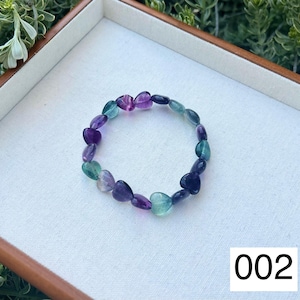 May include: A heart-shaped gemstone bracelet with purple, green, and blue stones. The bracelet is on a white surface with the number 002 printed on it.