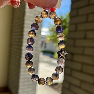 May include: A beaded bracelet featuring round, translucent beads in shades of gold and purple. A silver-colored star charm accents the bracelet. The bracelet is held up against a bright sky.