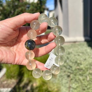 May include: A stretch bracelet composed of translucent, pale green spherical beads and one single dark, mottled blue and green spherical bead. A small white rectangular tag attached to the bracelet features the handwritten text 26-174.