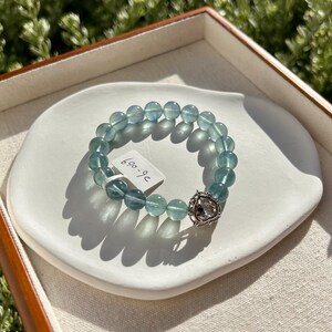 May include: A blue-green beaded bracelet with a decorative silver clasp. The bracelet is displayed on a white, oval-shaped dish inside a wooden tray. A small tag with the numbers "690-96" is attached to the bracelet.