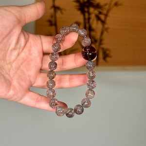 May include: A beaded bracelet featuring small, speckled, translucent beads and a larger, dark brown, spherical bead. The bracelet is held up against a neutral background, showcasing its natural colors and textures. The beads appear to be approximately 0.5 inches in diameter.