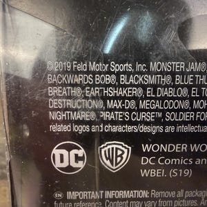 May include: A close-up of a product package with the DC Comics logo and the WB shield. The text includes "WONDER WOMAN" and a list of monster truck names. The package is black with white text.