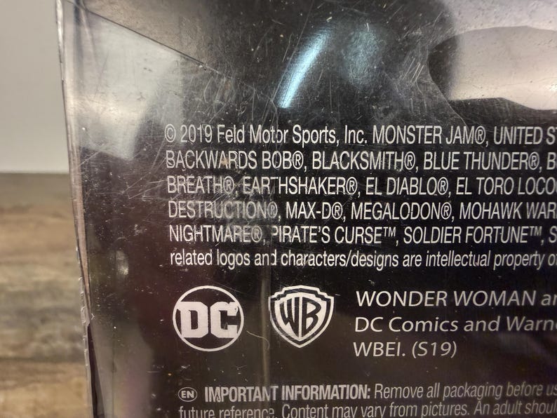 May include: A close-up of a product package with the DC Comics logo and the WB shield. The text includes "WONDER WOMAN" and a list of monster truck names. The package is black with white text.