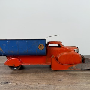 May include: Vintage toy truck in red and blue. The truck features a blue cargo bed, a red cab, and a front blade. The toy has a distressed appearance, suggesting it is an antique.