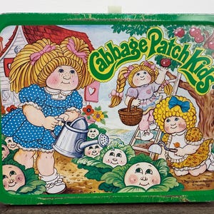 May include: Green metal lunchbox featuring a colorful illustration of Cabbage Patch Kids. The design includes several Cabbage Patch Kids, a house, and the words "Cabbage Patch Kids" in a green, stylized font. The lunchbox has a white handle.