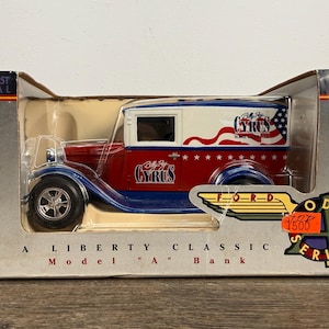 May include: Die-cast metal Liberty Classic Model A Bank. The red, white, and blue Ford model features the text "Billy Ray Cyrus" and a patriotic design. The model is in its original packaging.