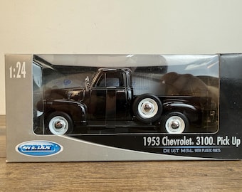 以下が含まれることがあります： 1:24スケールの箱に入った黒い1953年型シボレー3100ピックアップトラックのダイキャストモデル。車両には白いホイール、側面にマウントされたスペアタイヤ、シルバーのトリムが施されています。パッケージにはWellyブランドのロゴとモデル名が明記されています。