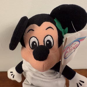 May include: A plush Mickey Mouse toy wearing a white robe and a green leaf headband. The toy has a black nose and large black eyes. The toy is holding a tag that says "Disney Store".