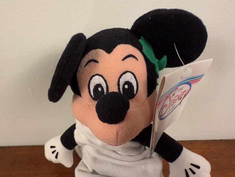 May include: A plush Mickey Mouse toy wearing a white robe and a green leaf headband. The toy has a black nose and large black eyes. The toy is holding a tag that says "Disney Store".