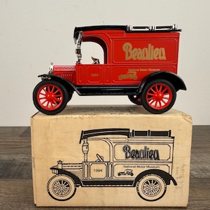 May include: A red and black vintage toy truck with the word "Beaulieu" in gold lettering. The toy truck is sitting on top of a cardboard box with a black and white illustration of the truck. The wheels are red.