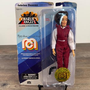 May include: A Sabrina Duncan action figure from Charlie's Angels, packaged in a clear plastic case. The figure is wearing a red jumpsuit and white shirt. The packaging includes the text "CLASSIC 8 INCH FIGURE" and "14 POINT ARTICULATION".