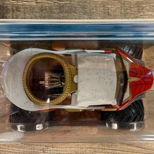 May include: A toy monster truck with a white body, red and gold accents, and large black tyres. The truck features a gold roll cage and a red spiderweb design on the front. It is packaged in a clear plastic container.