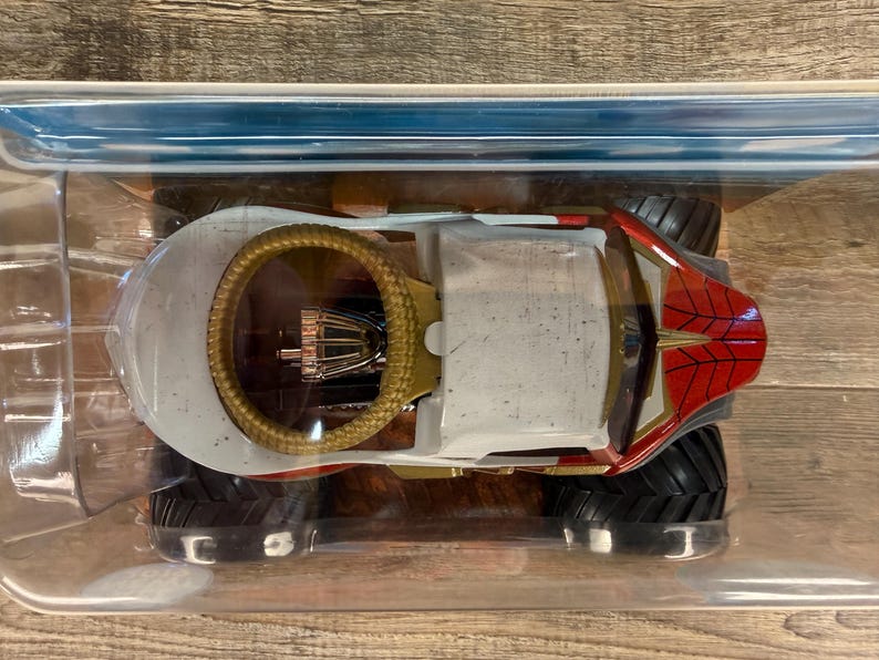 May include: A toy monster truck with a white body, red and gold accents, and large black tyres. The truck features a gold roll cage and a red spiderweb design on the front. It is packaged in a clear plastic container.