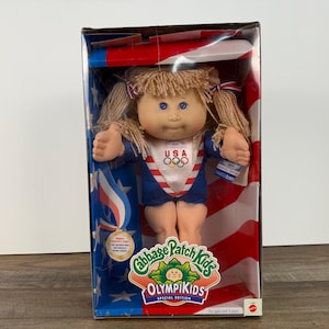 May include: A Cabbage Patch Kids OlympiKids Special Edition doll in a clear box. The doll has blonde pigtails and is wearing a blue and white USA uniform. The box features a red, white, and blue color scheme with a medal and the Cabbage Patch Kids logo.