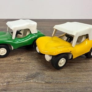 May include: Two toy dune buggies, one green and one yellow, with white tops and black tires. The buggies have a simple design with visible steering wheels and white seats. They are set on a wooden surface.